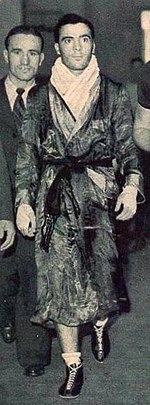 Photographie en noir et blanc d'un boxeur marchant, habillé d'un peignoir, serviette autour du cou et mains bandées.